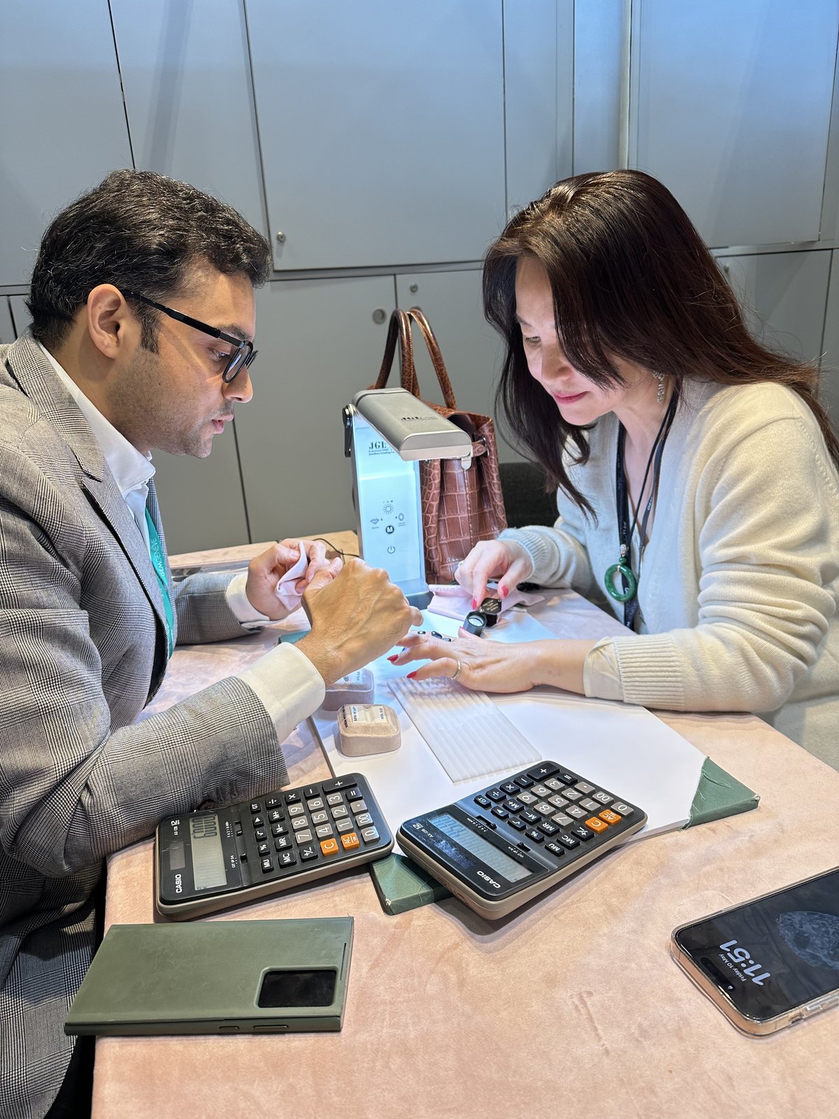 Kehan Li at a GemGenève table examining a single stone, with calculators, gem tools, and stone papers laid out across the surface.